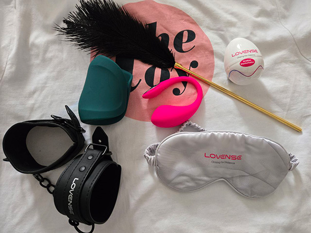 All six items from the Lovense Lover Set displayed on white bedding: a coral-pink Lush 4 egg vibrator, dark Gush 2 penis massager, translucent Kraken masturbator egg, black satin blindfold, black faux-leather adjustable wrist cuffs, and a black-handled feather tickler