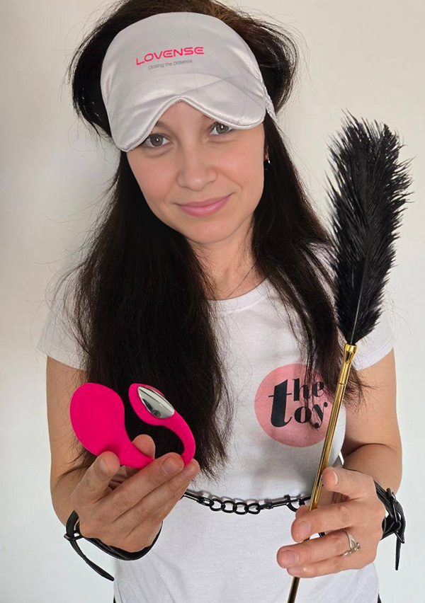 Amie Dawson holding in one hand Lovense Lush 4 and in the other the feather from the set. The blindfold on her head and demonstrating the handcufs