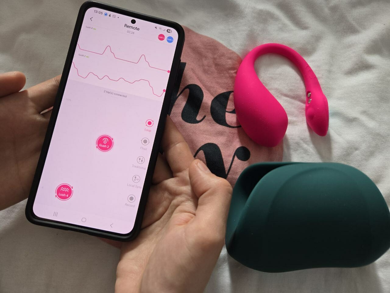 A person's hands—one holding a smartphone displaying the Lovense Remote app with two devices connected (Lush 4 and Gush 2 shown on screen), the other hand loosely holding a black-handled feather tickler against a dimly lit bedroom background.