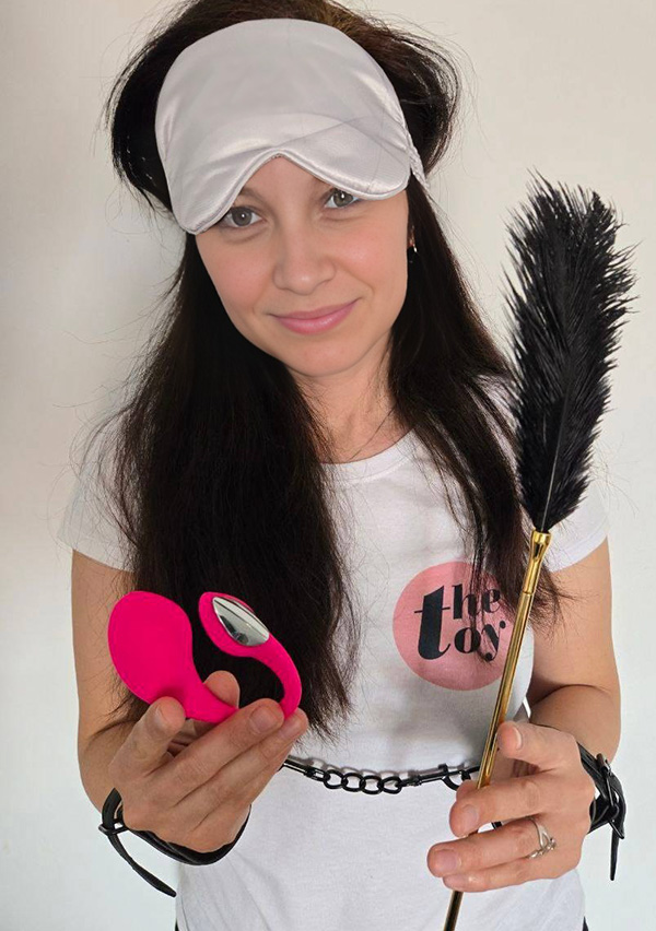 Amie Dawson Ph. D. with a blindfold on her forehead, Lovense Lush 4 in her left hand, a feather tickler in her right hand, and handcuffs on both wrists.