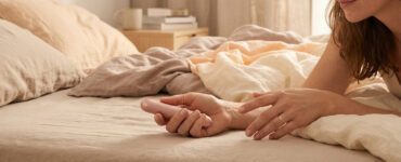 Soft editorial photo of a hand near a small vibrator on neutral bedding, suggesting a thoughtful comparison between toy stimulation and touch.