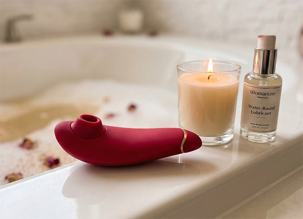 Womanizer Premium 2 resting on a white bathtub edge beside a candle and water-based lubricant, illustrating bath-time use.