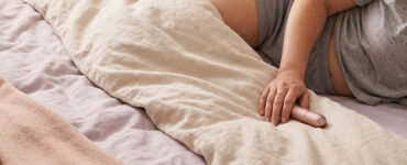 Soft editorial photo of a hand resting with a vibrator on neutral bedding in warm light, suggesting a paused intimate moment rather than active use.