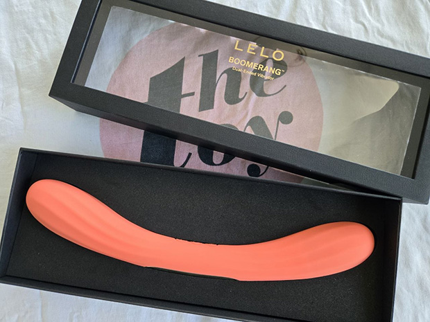 The LELO Boomerang double-ended vibrator in coral orange, displayed in its full U-shaped curve showing both ribbed insertable ends connected by a solid middle section with three control buttons in its luxury box