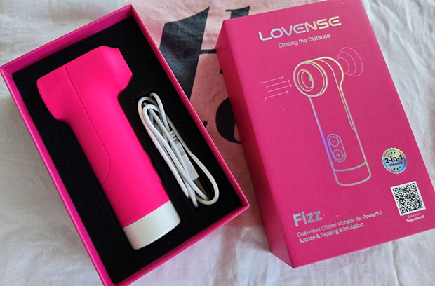 Lovense Fizz clitoral suction and tapping vibrator displayed alongside its packaging and USB charging cable, with the LED base light illuminated in soft pink