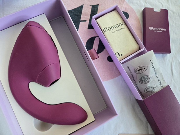 Everything included in the Womanizer Next Duo box: the dual-stimulation device, a cotton drawstring storage pouch, an extra size-M silicone stimulation head, a magnetic USB charging cable, and the quick-start guide.